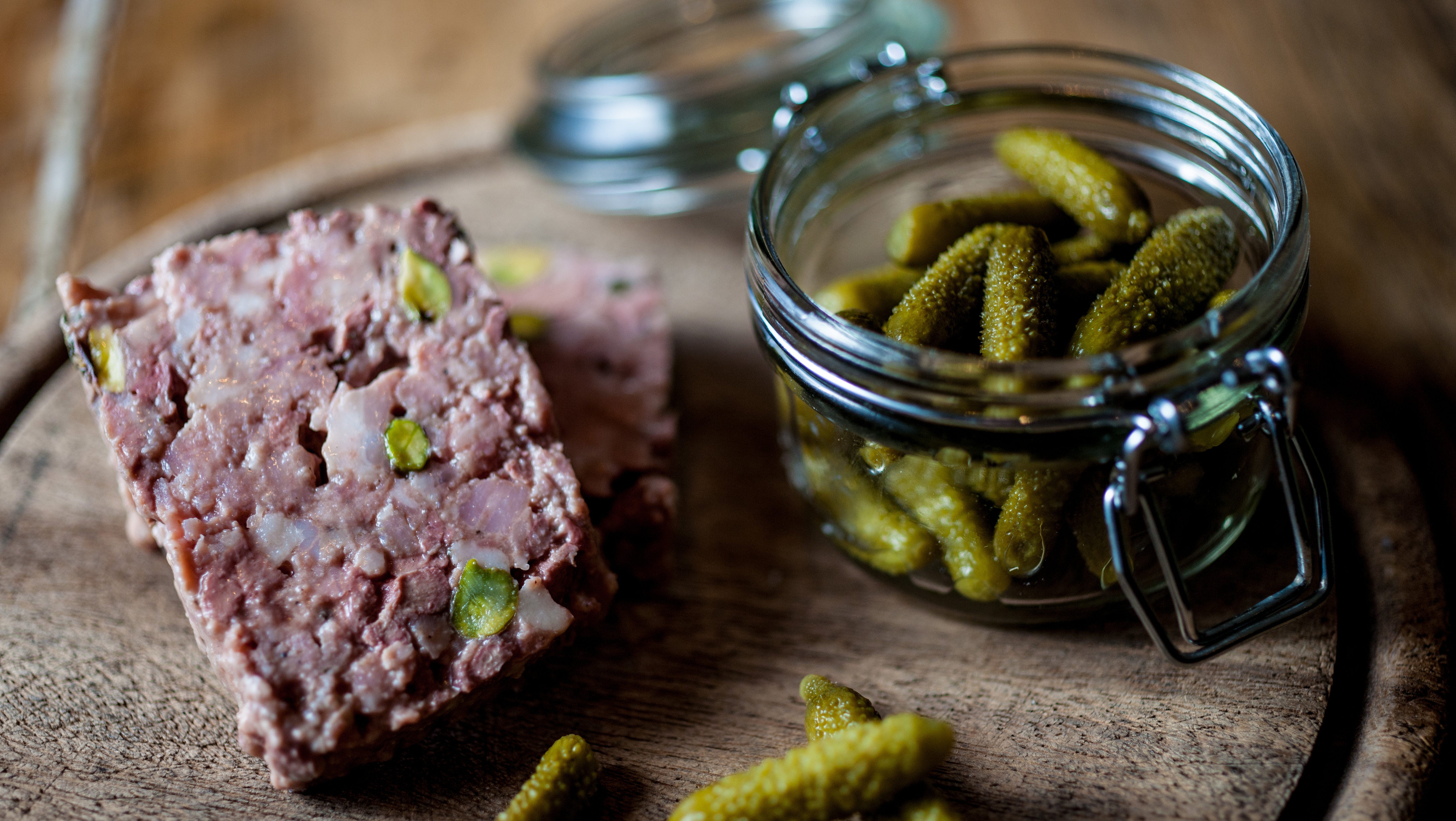 Close-up of sliced meat terrine with pistachios next to a jar of pickled gherkins.